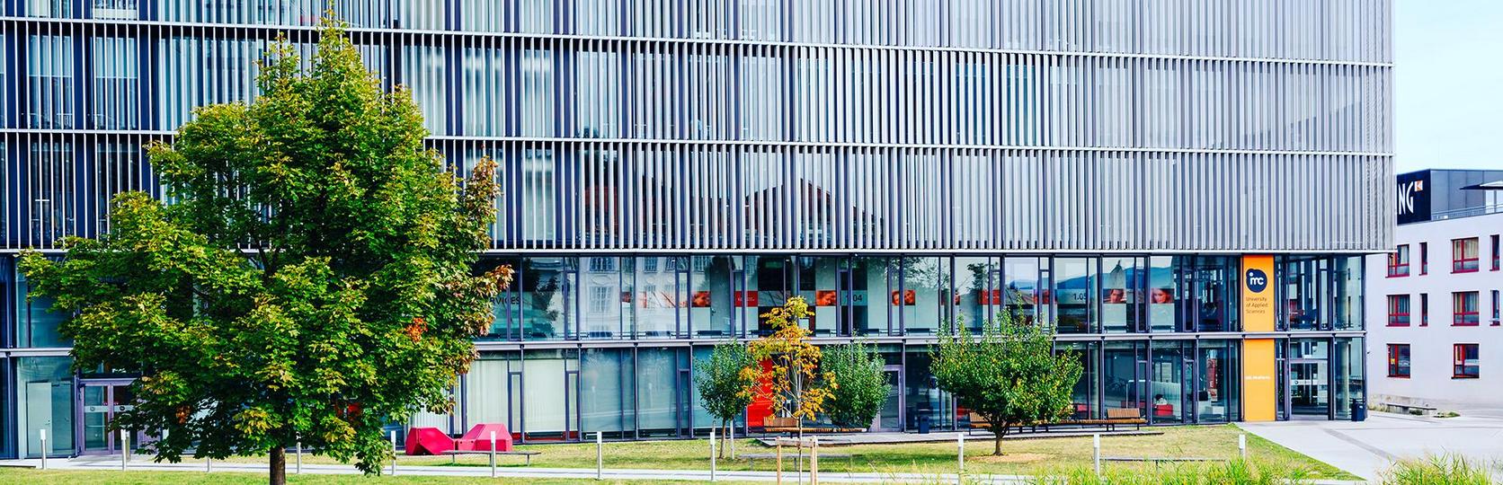 Modernes Gebäude mit Glasfassade und externen Jalousien, umgeben von Grünflächen mit einem Radfahrer im Vordergrund.