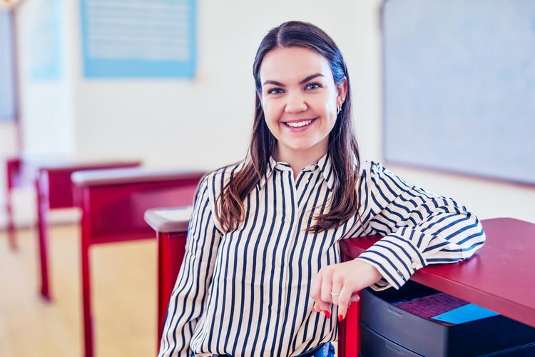 Person sitzt alleine an einem Schreibtisch in einem Klassenzimmer mit roten Stühlen und Tischen sowie einer Tafel im Hintergrund.
