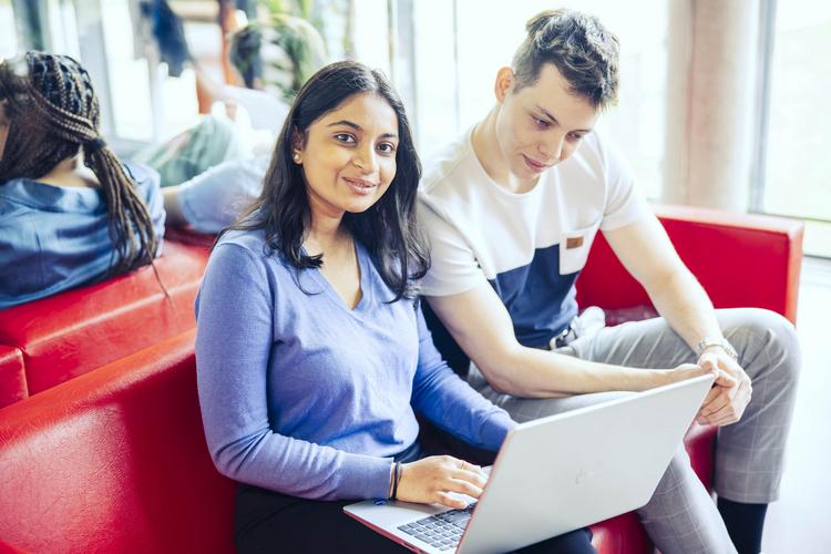 Zwei Personen sitzen nebeneinander auf einer roten Couch. Das Mädchen grinst in die Kamera und hat einen Laptop auf dem Schoß. Der Junge starrt gespannt auf den Bildschirm. Im Hintergrund sitzen weitere Personen mit dem Rücken zur Kamera.