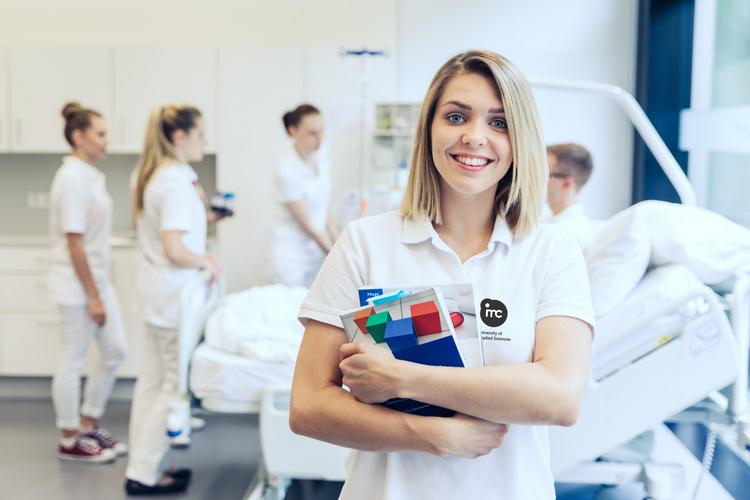Eine Gruppe von Personen in medizinischer Kleidung steht in einem unserer Health Labs, in denen ein Krankenhausumfeld simuliert wird. Eine weibliche Person im Vordergrund hält Lehrmaterialien und lächelt in die Kamera.
