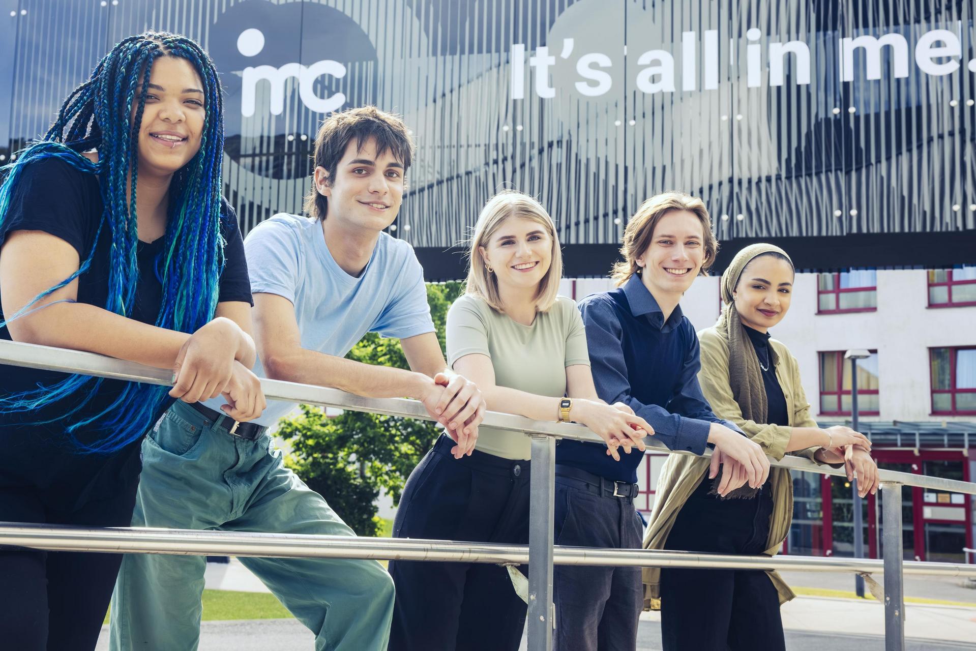 A group of five students lean smiling against an outdoor railing, with an IMC building in the background