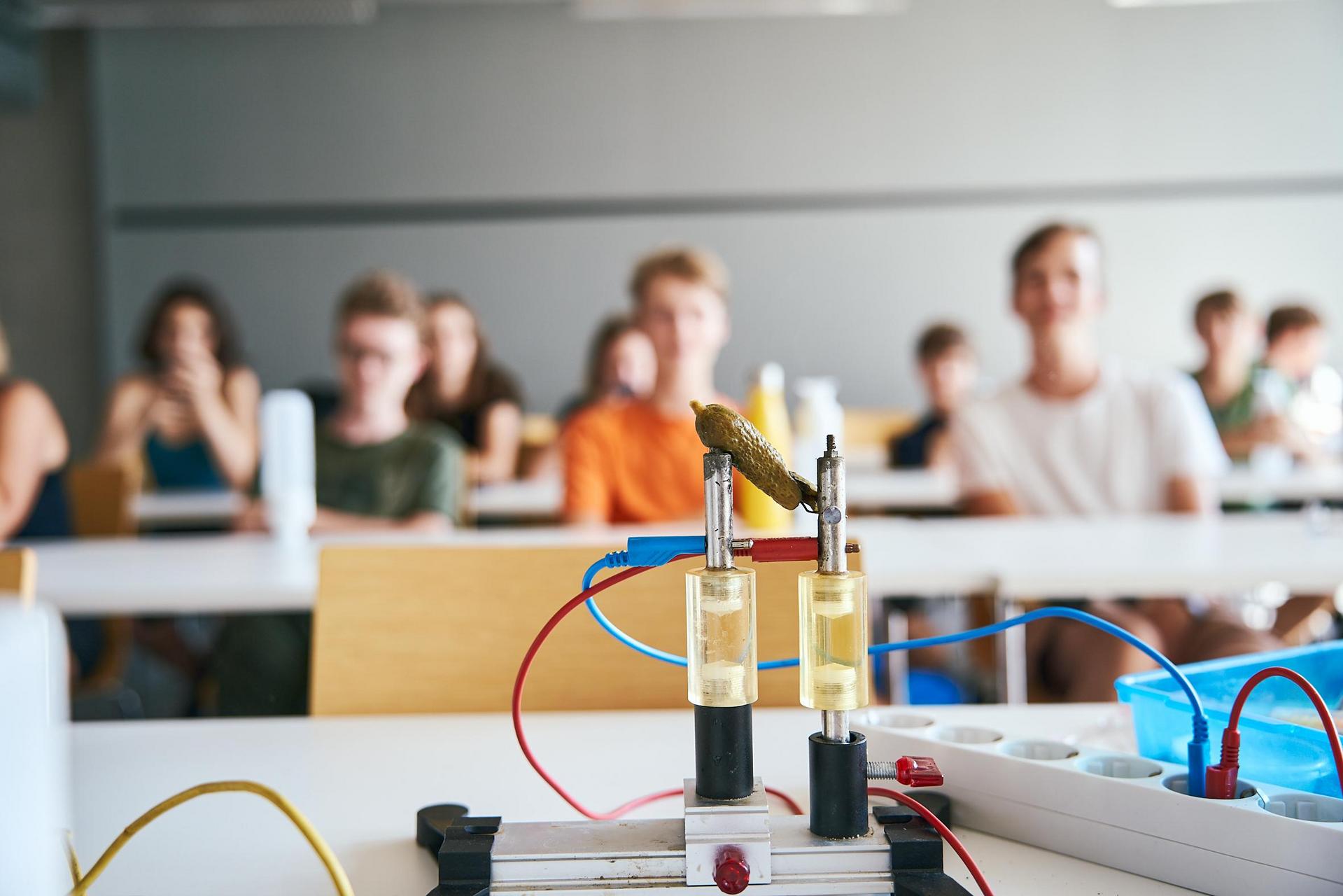 Ein Klassenzimmer mit unscharfen Schülern im Hintergrund und einem Fokus auf elektronische Bauteile und Verkabelungen im Vordergrund