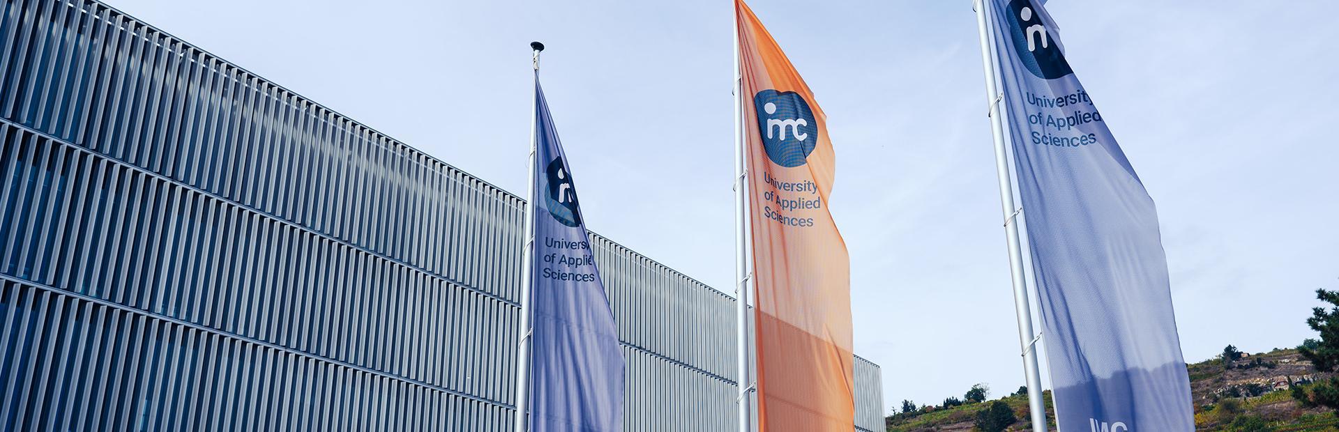 Zwei graue und eine orange Fahne am IMC Campus Krems, jeweils mit dem IMC Krems Logo. Gebäude Trakt G ist im Hintergrund zu sehen.