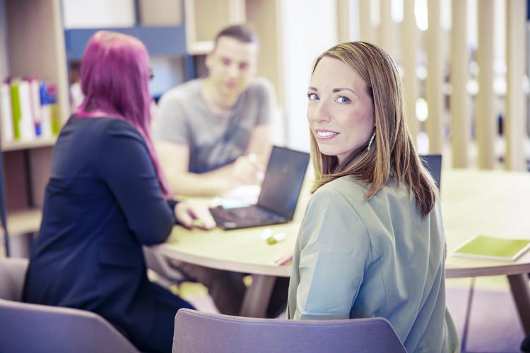 Drei Studierende sitzen um einen kleinen runden Tisch in einem Lernbereich einer österreichischen Hochschule. Einer benutzt einen Laptop und auf dem Tisch liegen Bücher, was auf eine akademische Umgebung hinweist.