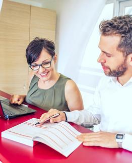 Zwei Personen arbeiten an einem Schreibtisch, eine benutzt einen Laptop und die andere schreibt in ein Notizbuch.