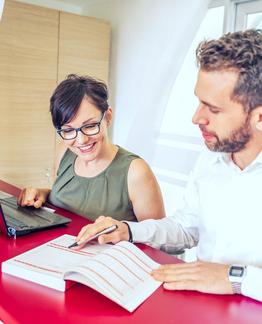 Two individuals working at a desk, one using a laptop and the other writing in a notebook.