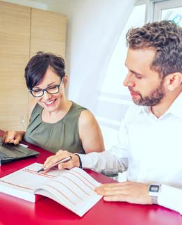 Two individuals working at a desk, one using a laptop and the other writing in a notebook.