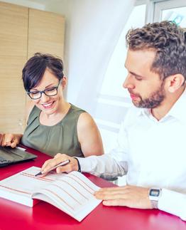 Zwei Personen arbeiten an einem Schreibtisch, eine benutzt einen Laptop und die andere schreibt in ein Notizbuch.
