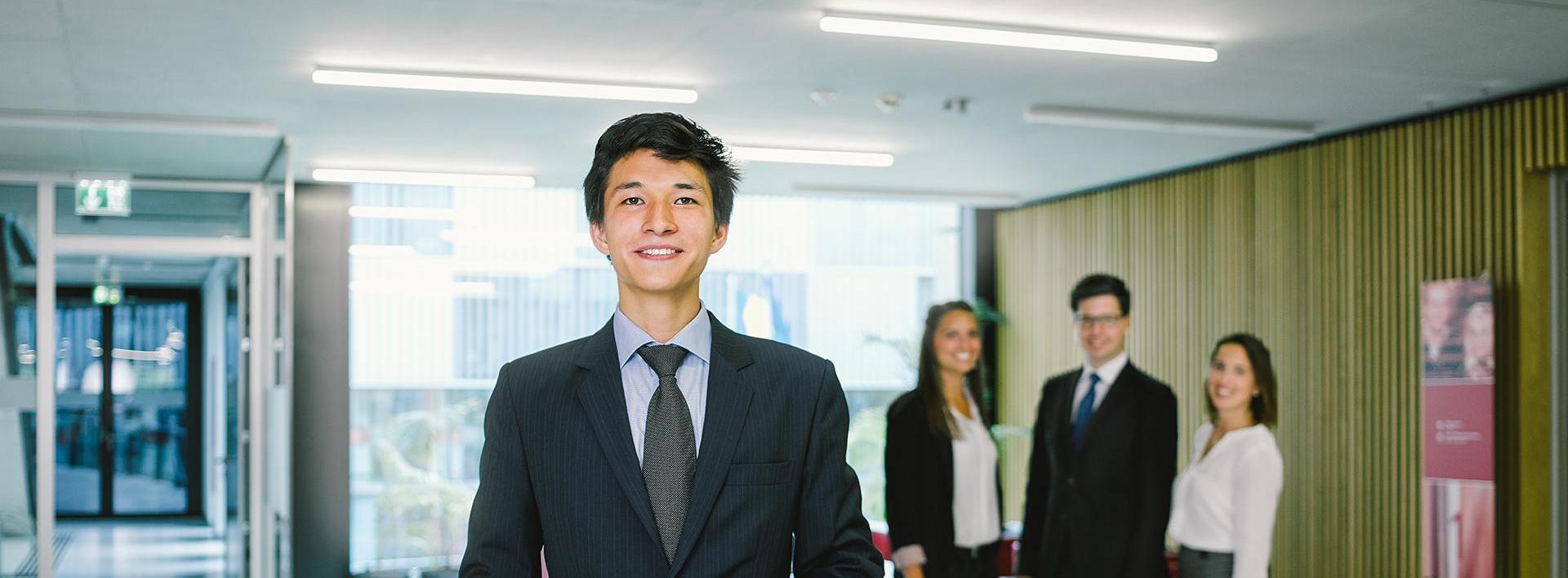Geschäftsmann mit verschränkten Händen steht im Büroflur, Kollegen im Hintergrund.