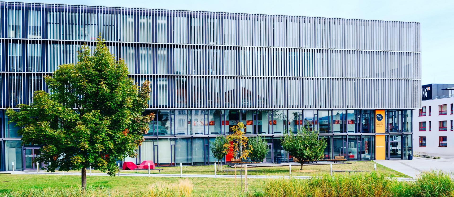 Modernes Bürogebäude mit Glasfassade und Metalllamellen, umgeben von Grünflächen und einem kleinen Graben im Vordergrund.