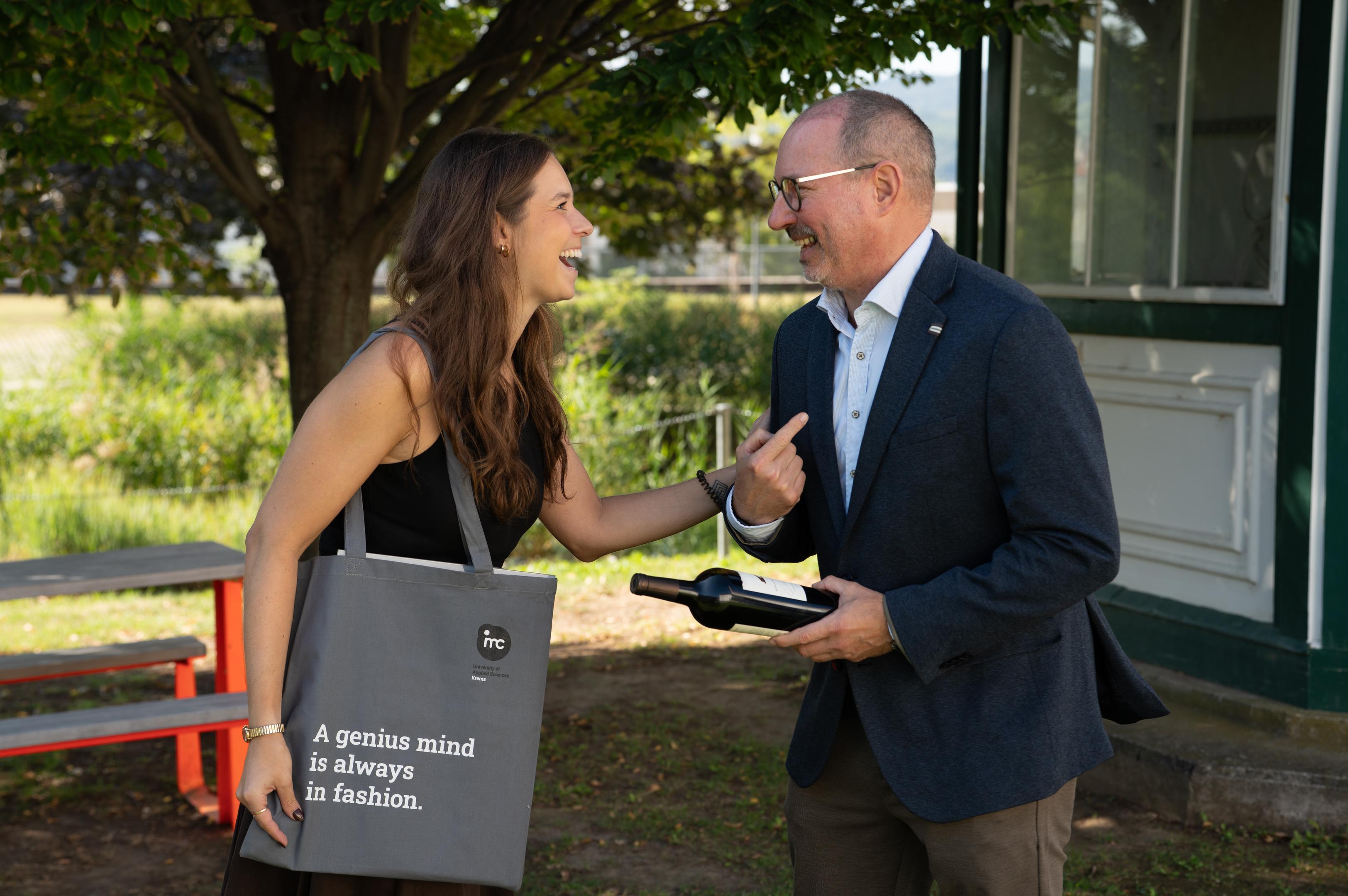 Frau und Mann lachen, sie hält eine IMC-Tasche, er eine Flasche Wein.