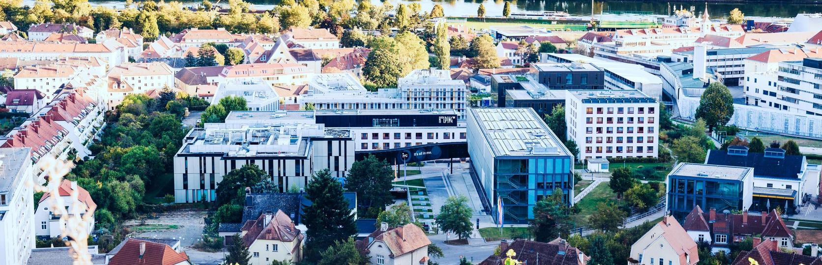 Stadtbild mit modernen und traditionellen Gebäuden, umgeben von grüner Landschaft und einem Fluss im Hintergrund.