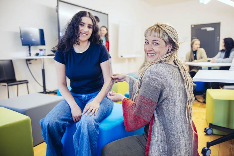 Zwei Personen sitzen sich in einem hellen Seminarraum am IMC Krems gegenüber. Eine Person mit langen blonden Haaren trägt eine rote Jacke und berührt den Arm einer anderen Person, die auf einem blauen Gymnastikball sitzt und ein dunkelblaues T-Shirt trägt. Im Hintergrund sind weitere Personen und ein großer Bildschirm zu sehen.