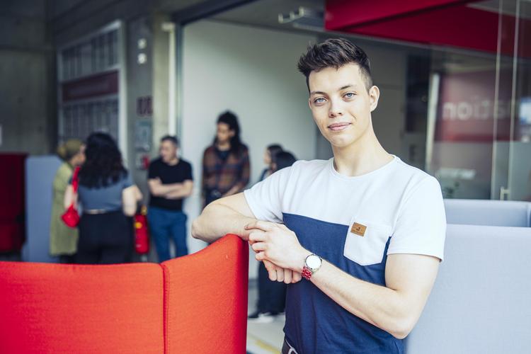Ein junger Mann steht lässig an einem Sessel und hat seinen Ellbogen auf der Lehne abgestützt. Im Hintergrund steht unscharf eine Gruppe von Personen, die sich miteinander unterhalten.
