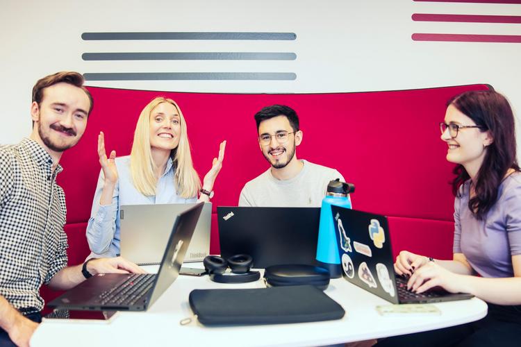 Vier Personen arbeiten zusammen an einem Tisch mit Laptops in einer modernen Umgebung mit roter Couch. Die Szene findet in einer unserer IMC Lounges statt.