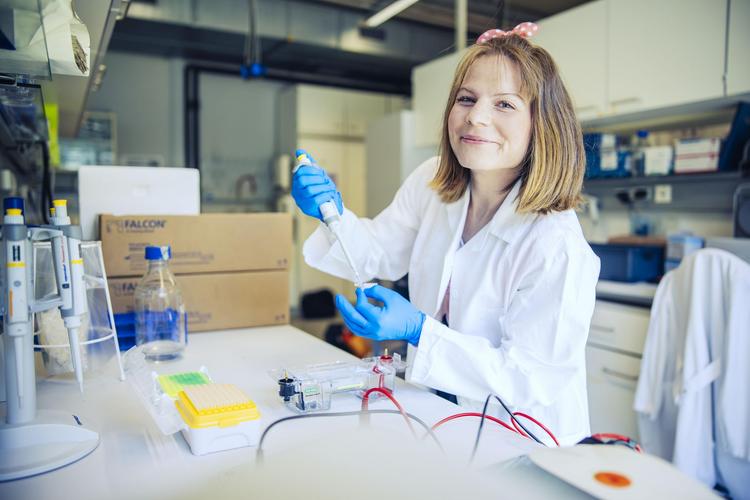 Eine Studentin im Labor trägt einen Laborkittel und Gummihandschuhe. Sie lächelt und verwendet eine Pipette, um eine Flüssigkeit in ein kleines Röhrchen zu übertragen. Im Hintergrund sind Laborausrüstung und Kartons sichtbar.