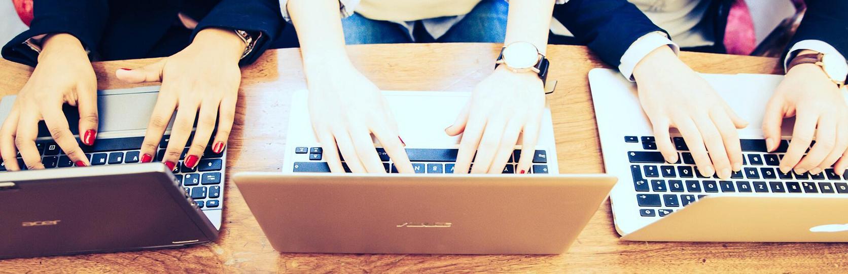 Drei Personen arbeiten an Laptops auf einem Holztisch, Fokus auf den Händen und Tastaturen.
