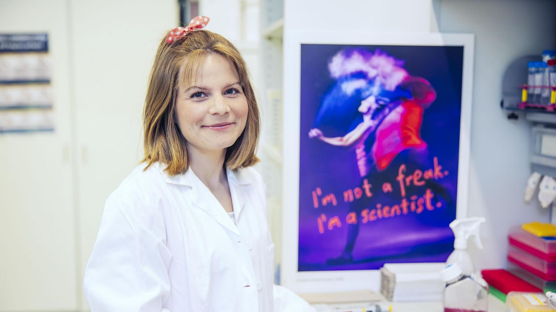 A person in a white lab coat stands in front of a poster with the inscription “On est fait pour s’entendre!”
