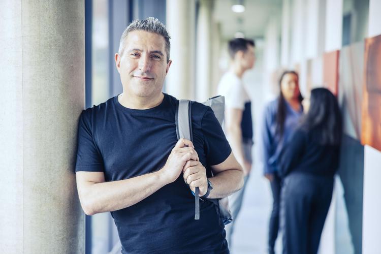 Ein Student steht lächelnd in einem Gang. Im Hintergrund sind drei weitere Studierende zu sehen, die miteinander sprechen. Die Szene findet auf dem Campus der Hochschule statt