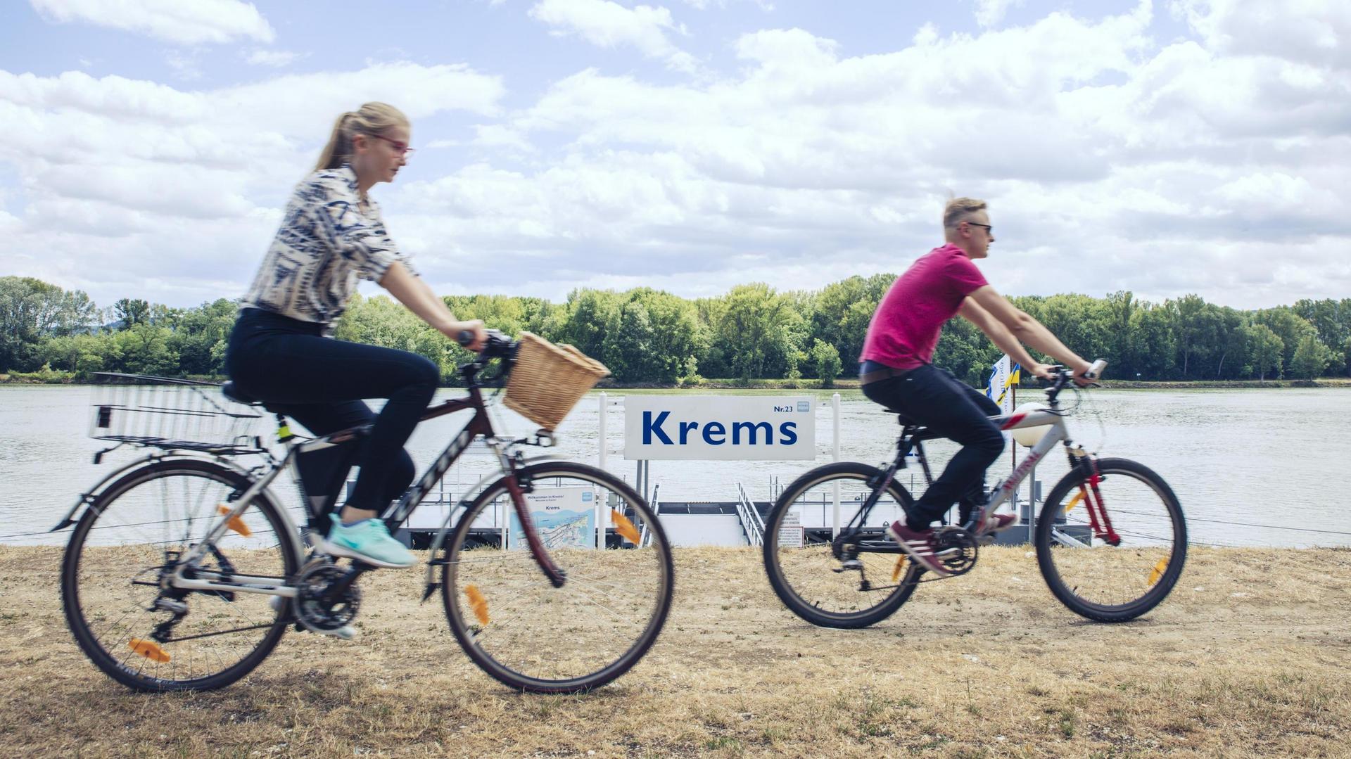 Two individuals riding bicycles along the river Danube with a sign displaying “Krems”.