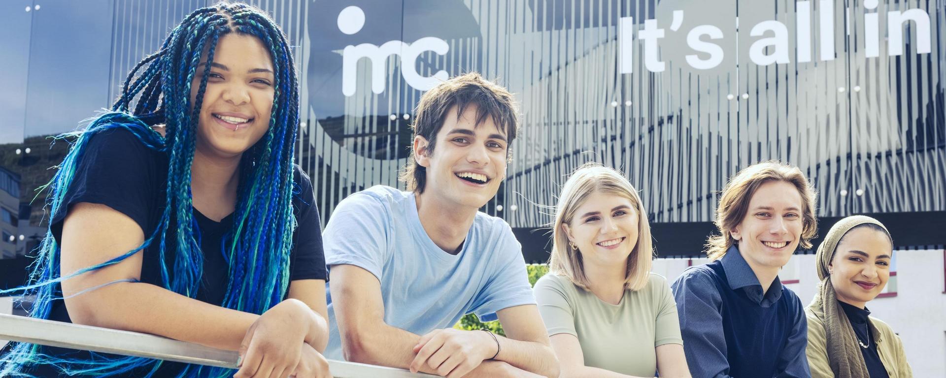 Gruppe von fünf jungen Menschen, die sich an einem Geländer vor einem modernen Gebäude mit reflektierenden Fenstern und der Aufschrift “it’s all in” lehnen.