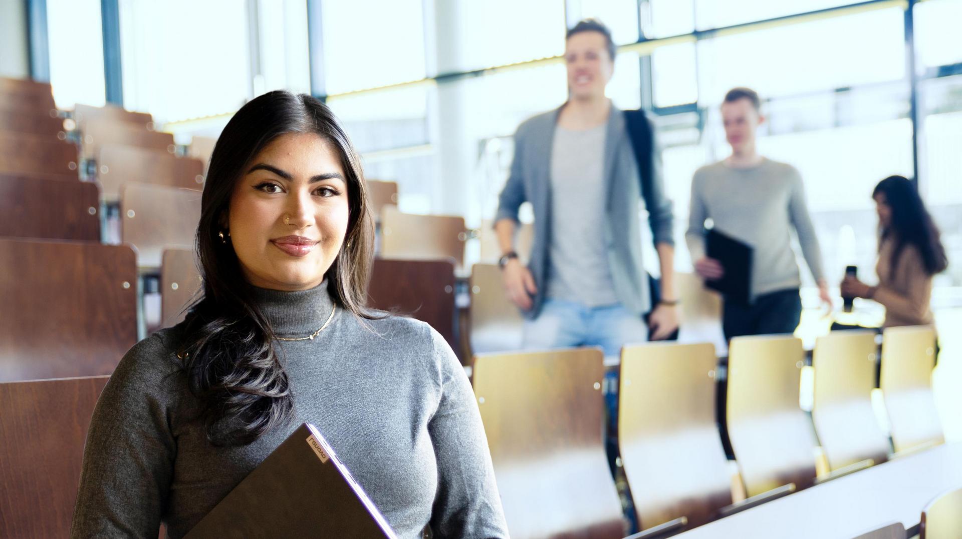 Studentin mit Buch steht im Hörsaal, während andere Personen im Hintergrund sitzen und gehen.