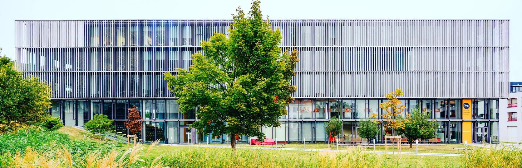 Modernes Bürogebäude mit Glasfassade und Metallverkleidung hinter einem Teich mit Schilf im Vordergrund.