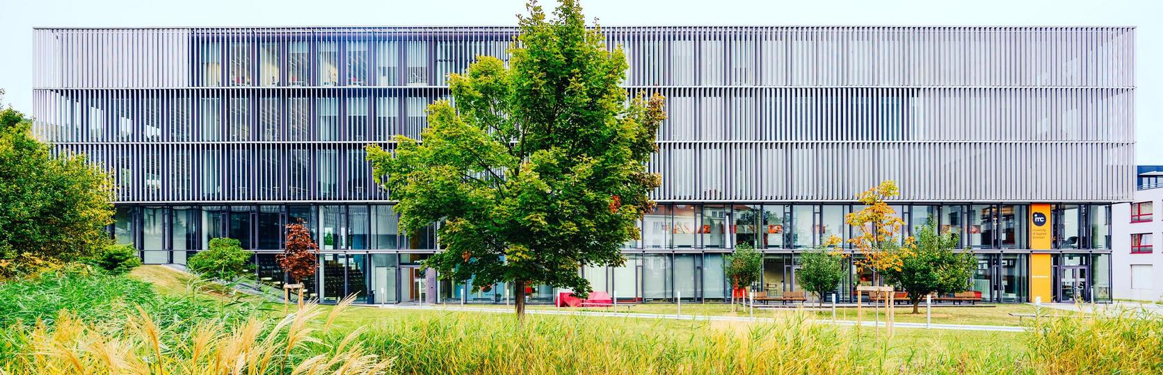 Modernes Bürogebäude mit Glasfassade und Metallverkleidung hinter einem Teich mit Schilf im Vordergrund.