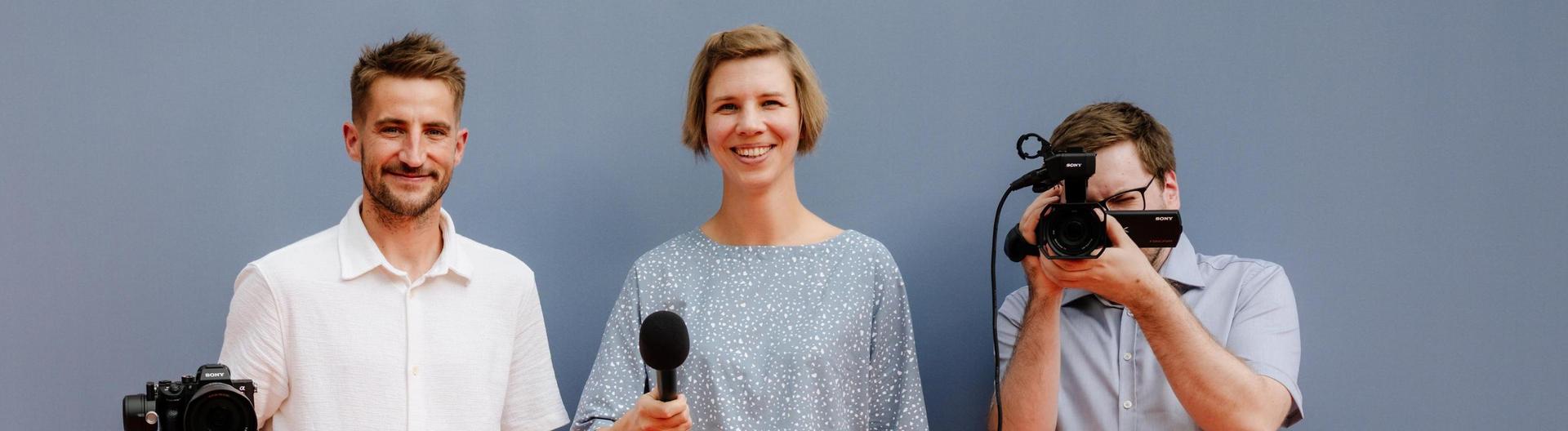 Drei Personen vor einer grauen Wand, von links nach rechts: eine Person mit einer Videokamera, eine Person mit einem Mikrofon und eine Person mit einer Fotokamera.