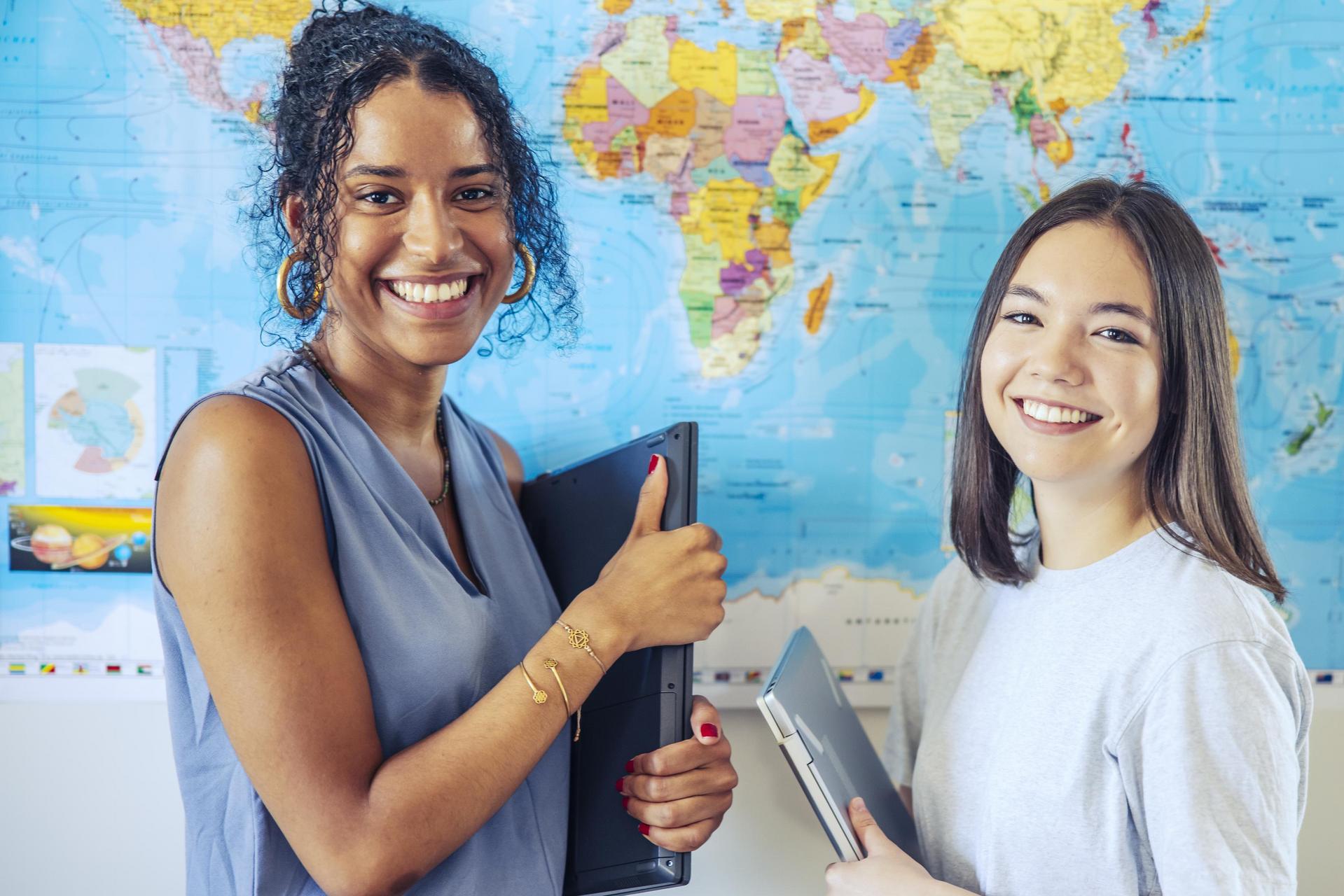 Two women stand in front of a world map and laugh into the camera. They are both holding a folder or notebook in the camera.