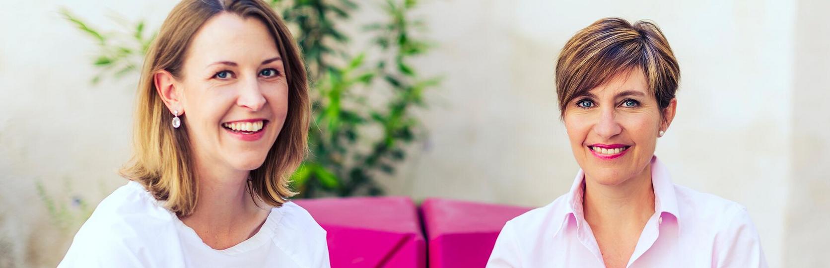 Zwei Personen sitzen nebeneinander vor einer hellrosa Couch und einer grünen Pflanze.
