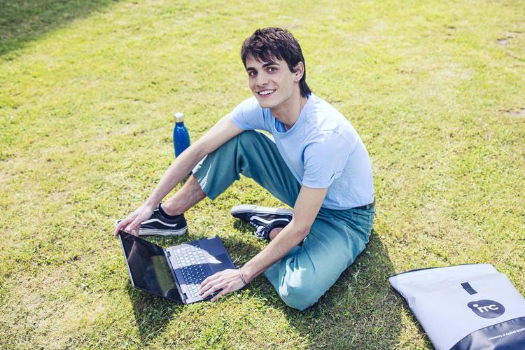 Ein junger Mann sitzt lächelnd auf einer Wiese und arbeitet an einem Laptop. Neben ihm steht eine blaue Trinkflasche, und ein Rucksack mit dem IMC Logo liegt auf dem Gras.