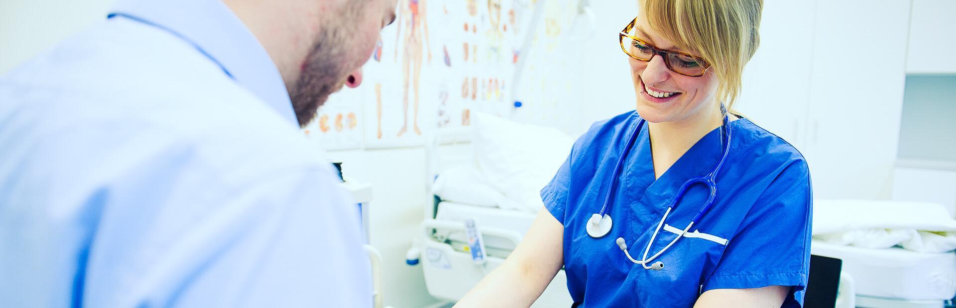Eine Person in blauen Pflegegewand untersucht in einer Übungssituation einen Patienten.