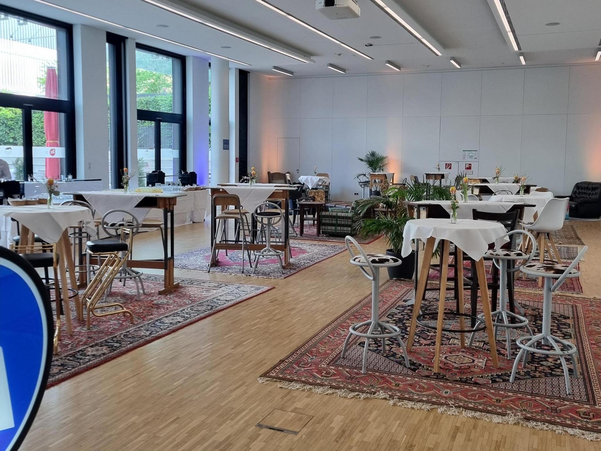 Modern gestalteter Innenraum mit Tischen und Stühlen, großen Fenstern und Pflanzen, beleuchtet durch Deckenleuchten