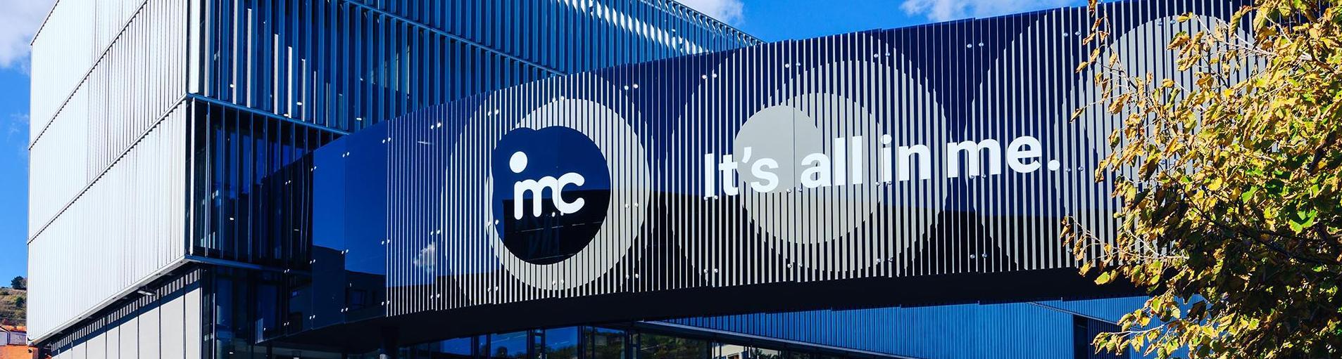 Modernes Gebäude mit blauer Fassade und dem Logo IMC sowie dem Slogan it’s all in me bei Tageslicht.