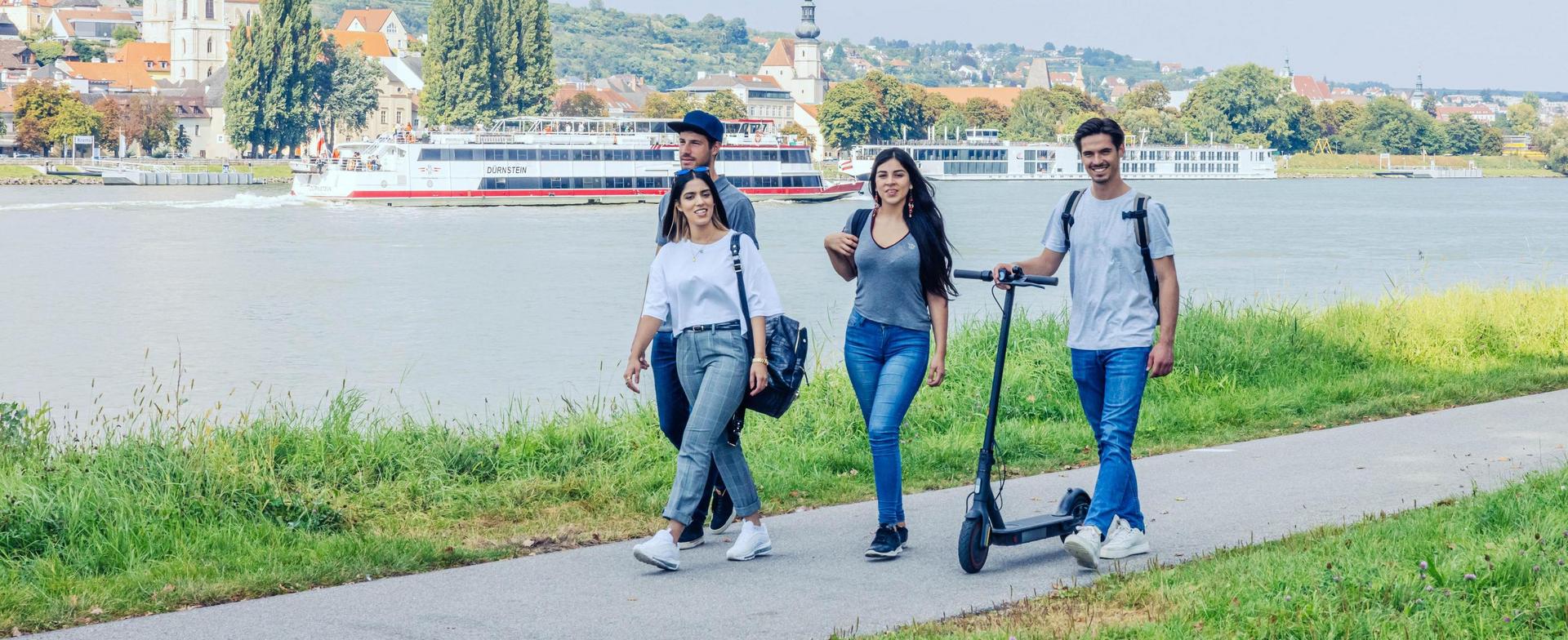 Four young adults are walking along a path next to the Danube, with one person pushing a black electric scooter. Krems an der Donau can be seen in the background