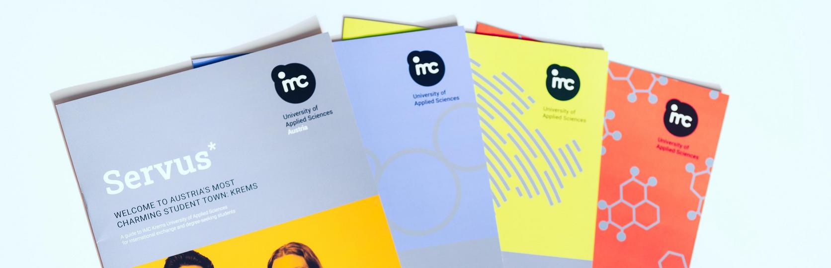 A stack of brochures with a focus on the top brochure featuring the IMC logo and the title Servus, promotional or informational material