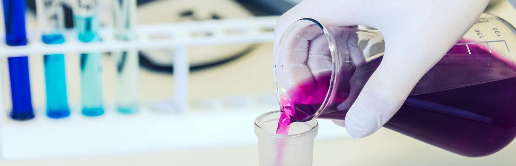 A lab assistant pours a purple liquid from a beaker into a test tube amidst other laboratory glassware and equipment.