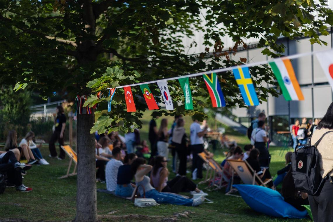 Flaggen von verschiedenen Ländern auf einer Schnur, im Hintergrund sitzen viele Leute im Gras und auf Sesseln