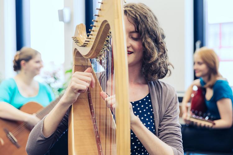 Eine Frau sitzt und spielt auf einer Harfe. Im Hintergrund sind rechts und links unscharf zwei weitere Frauen zu erkennen, die beide eine Gitarre in der Hand halten, sich gegenseitig anlächeln und ebenfalls spielen.