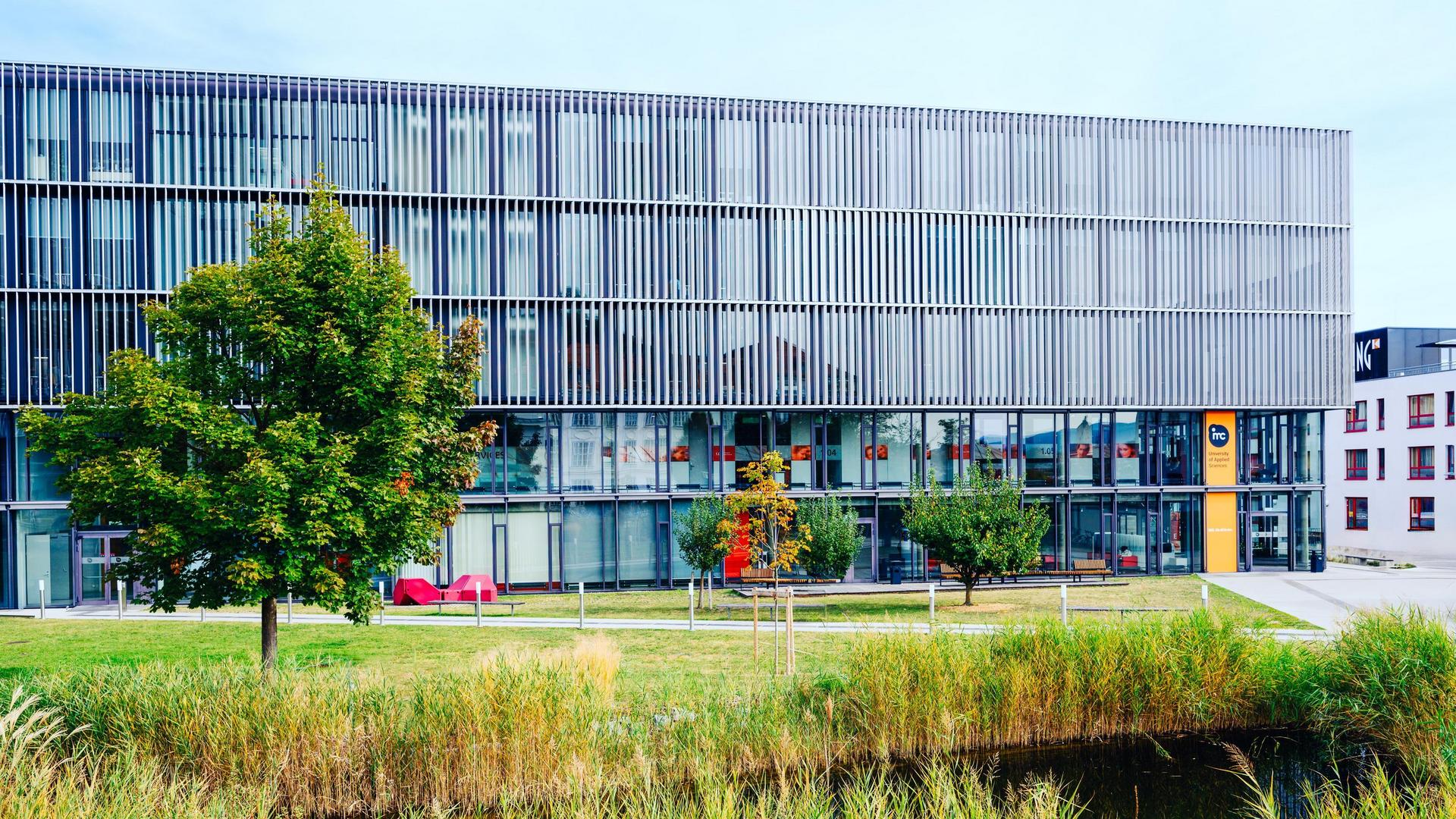 Modern office building with glass facade and metal slats, surrounded by green spaces and a small ditch in the foreground.