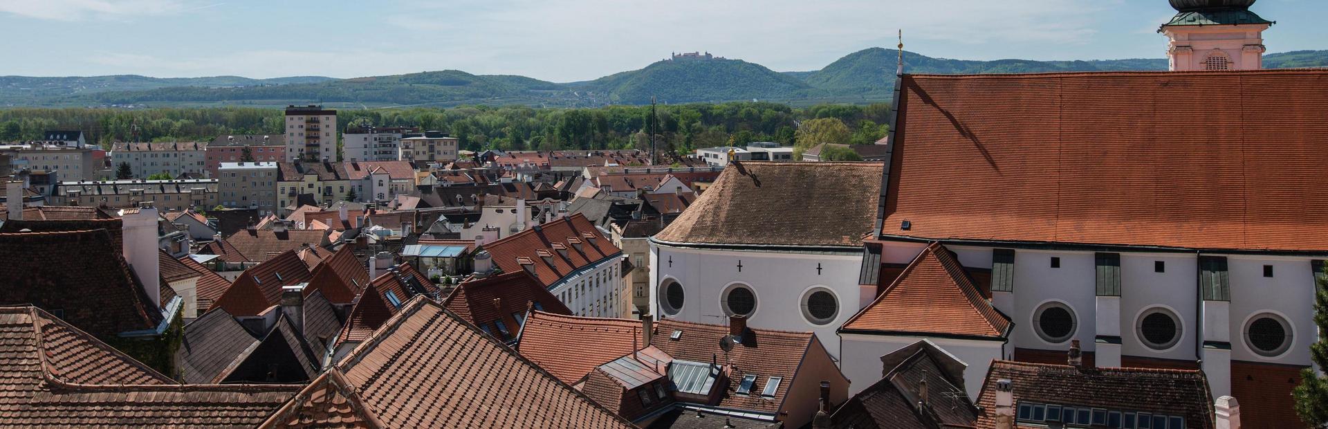 Cityscape of Krems with red tiled roofs and buildings against a wooded hill under a clear blue sky.
