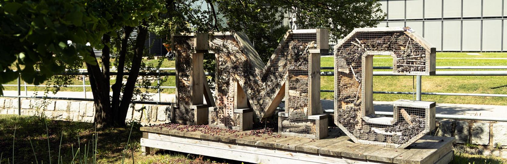 Large, three-dimensional IMC letters made of metal lattice overgrown with climbing plants on a wooden platform in front of a building and trees.