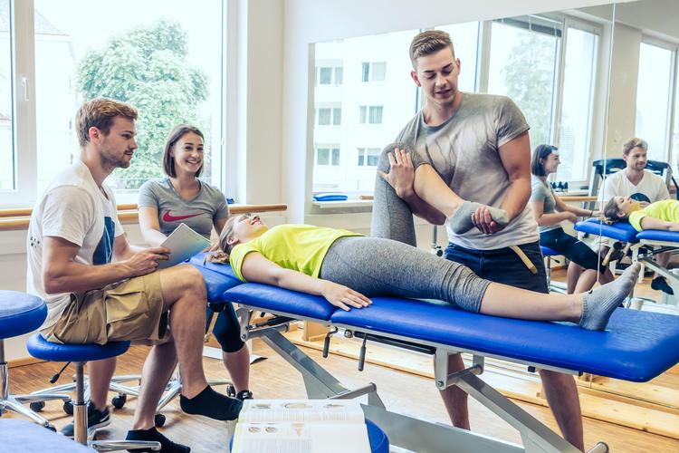 Es sind vier Personen zu sehen in einem hellen Physiotherapie-Übungsraum des IMC Krems. Eine Frau liegt auf einer blauen Liege während ein Mann eines ihrer Beine abwinkelt und dabei spricht. Am Kopfende der Liege sitzen zwei weitere Personen, die zuschauen - eine davon mit einem Block in der Hand, um sich Notizen zu machen.