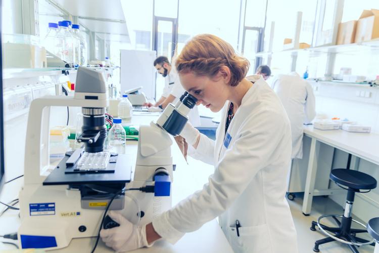 The photo shows a student in a white coat and white gloves in one of the IMC laboratories. She is using a microscope, which she is looking through, and is surrounded by laboratory equipment. A second person can be vaguely recognised in the background.