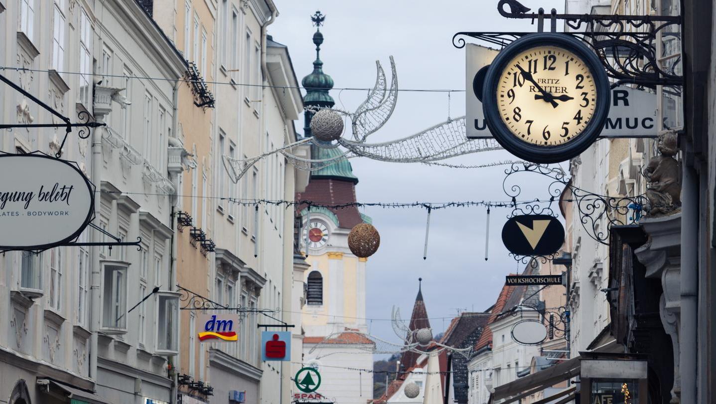 A bustling shopping street with historic buildings, street lamps, and a clock in the foreground conveying a busy city atmosphere