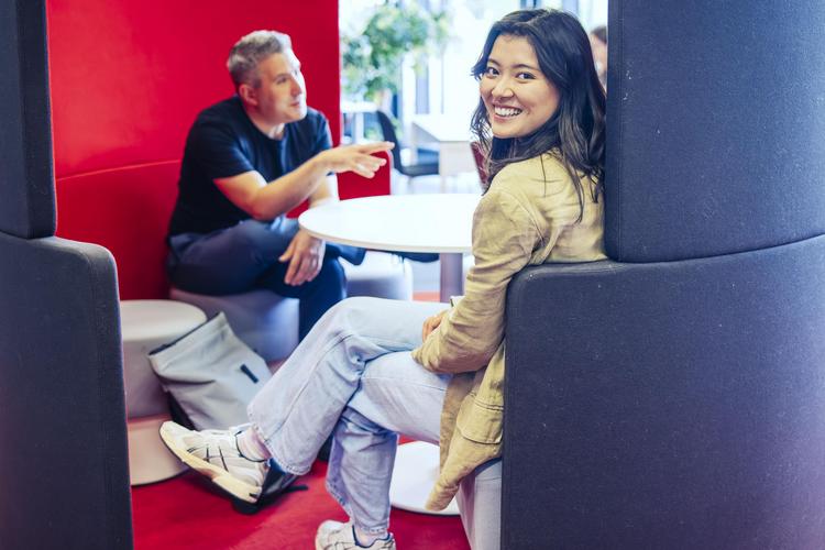 Zwei Personen sitzen an einem runden Tisch in der IMC Study Lounge gegenüber, umgeben von hohen Sitzgelegenheiten in einem Innenraum mit roten und grauen Farbakzenten. Eine Person lächelt über die Schulter in die Kamera und sitzt mit überschlagenen Beinen am Tisch. Die zweite Person ist unscharf auf der anderen Seite des Tisches zu erkennen und unterhält sich mit jemandem. 