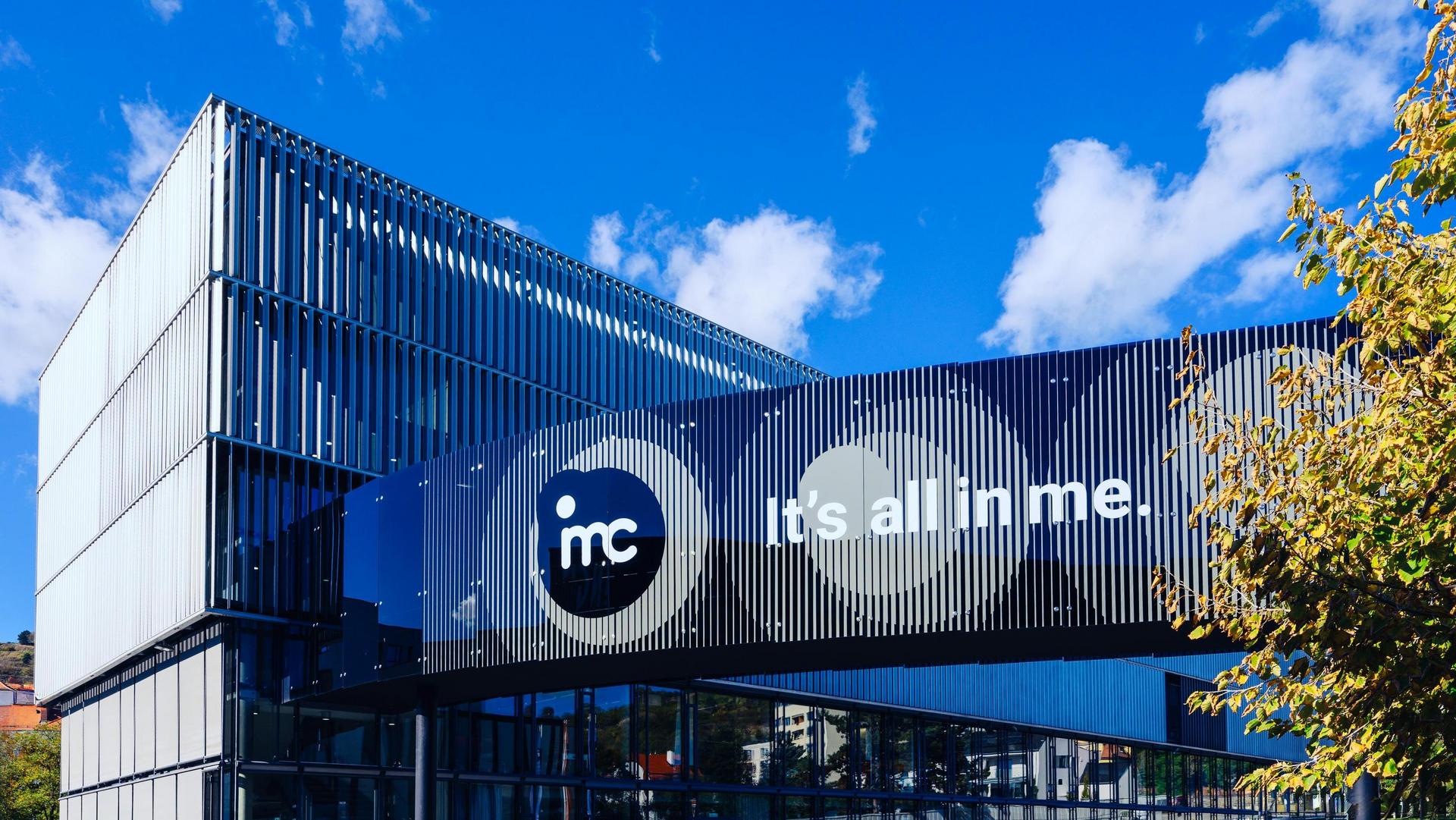 Modern building with blue and black panels, “mcc” logo and “it’s all in me” slogan under a clear blue sky.