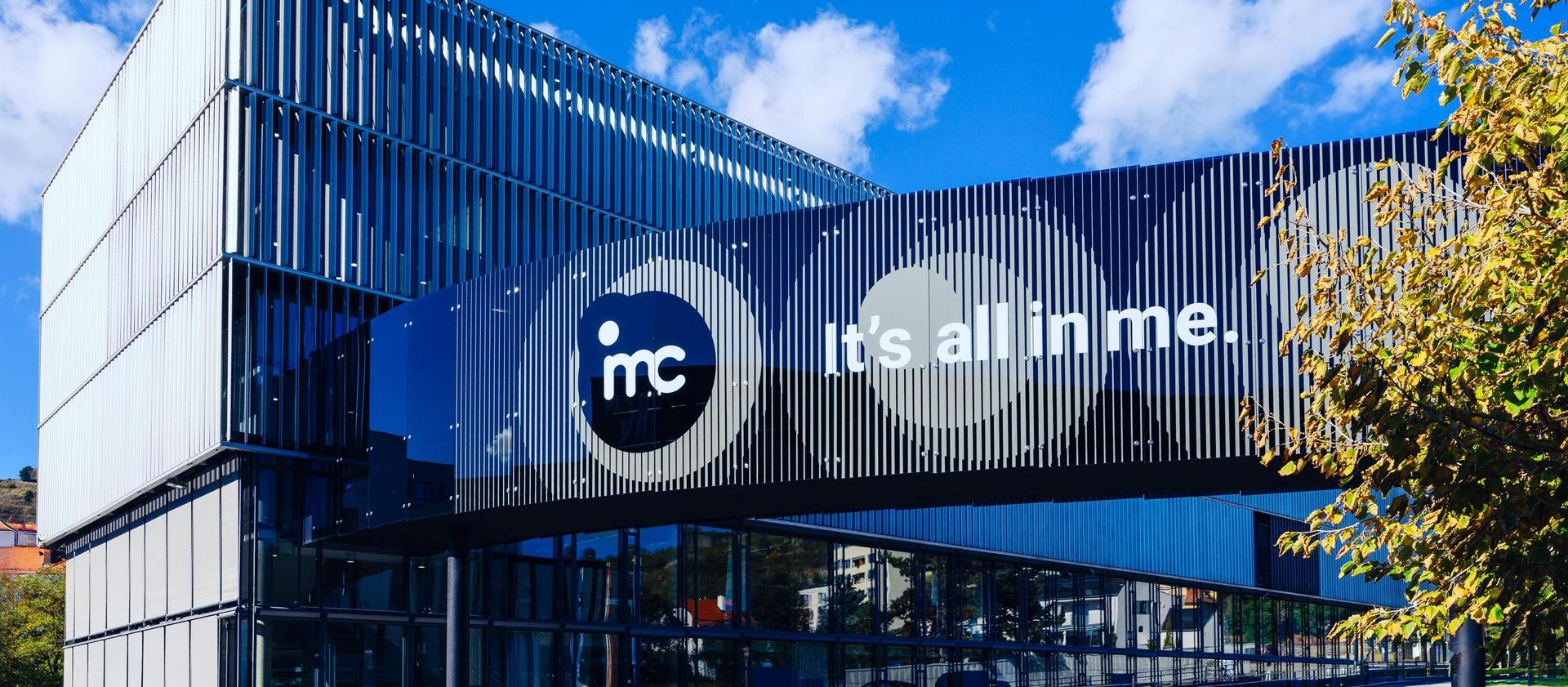 Modernes Gebäude mit blauen und schwarzen Paneelen, Logo “imc” und Slogan “it’s all in me” unter klarem blauem Himmel.