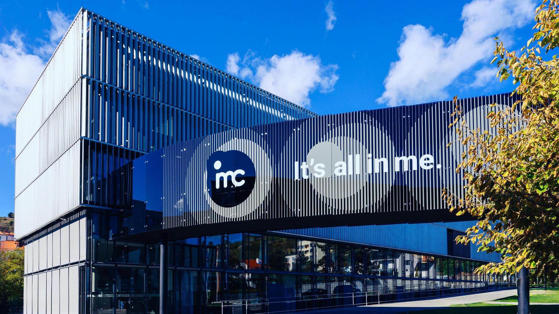 Modernes Gebäude mit blauen und schwarzen Paneelen, Logo “imc” und Slogan “it’s all in me” unter klarem blauem Himmel.